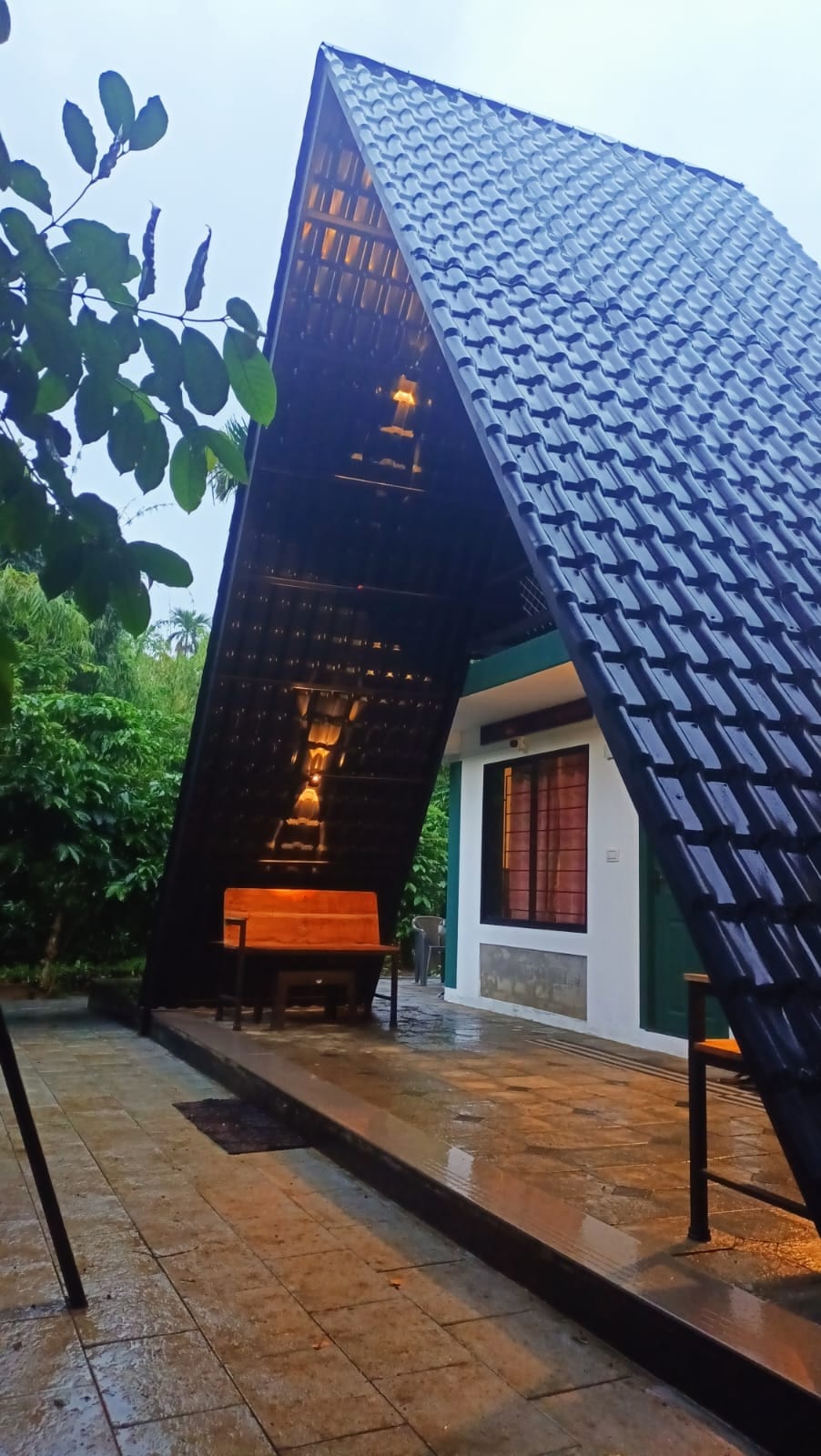 An A-frame sitout structure features a distinctive slanted roof, complemented by a wooden bench positioned beneath it. The entrance is framed by lush greenery, and the flooring consists of large stone tiles. Soft lighting highlights the architectural design during twilight.