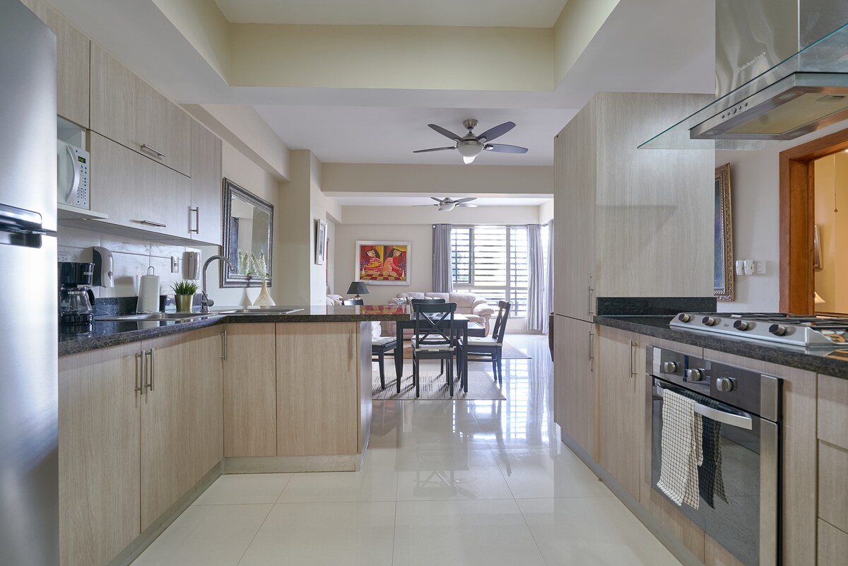 The kitchen space features modern cabinetry and appliances, including a refrigerator, oven, and stovetop. A dining area is visible in the background, highlighted by a ceiling fan. Natural light enters through large windows, illuminating the neutral-toned surfaces and tile floor.
