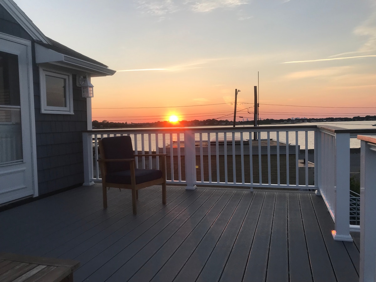 A deck is shown with a single chair positioned to view the horizon. The sun sets beyond the water, casting warm tones across the sky. The railing contrasts with the grey decking, providing a quiet space to enjoy the surroundings.