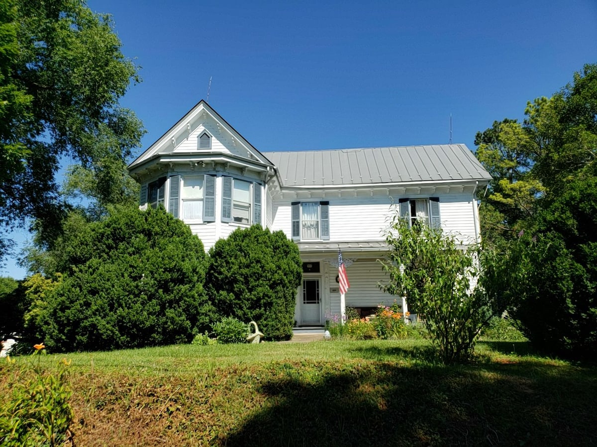 The exterior of a Victorian home is shown, featuring a white façade and a metal roof. Lush green bushes frame the entrance, and colorful flowers are visible in the garden. A flag is displayed near the front door. The clear blue sky enhances the charming setting.