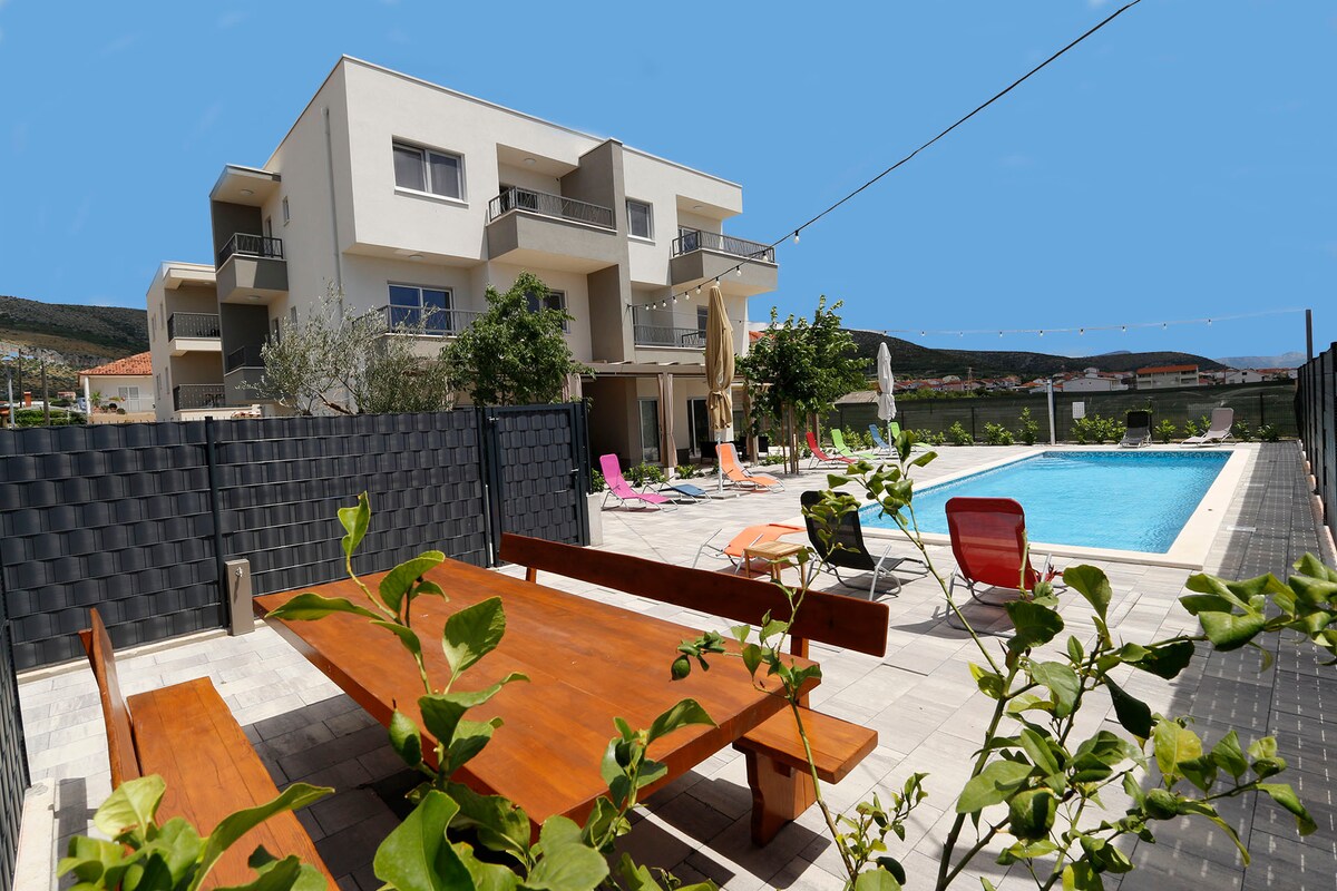 A modern outdoor area features a spacious wooden dining table surrounded by greenery. Colorful lounge chairs are arranged near the crystal-clear swimming pool, which is framed by a neat stone patio. The facility's contemporary buildings are visible in the background, complemented by lush hills.