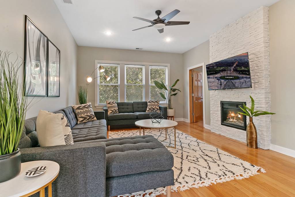 The spacious living room features a plush sectional sofa and a modern coffee table, arranged in a cozy seating area. A decorative area rug lies beneath, while plants add a touch of greenery. Natural light fills the room through three large windows, and a stylish fireplace serves as a focal point.