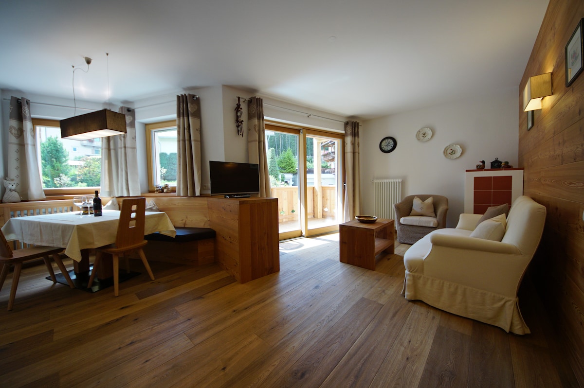 A welcoming living space features natural wooden flooring and large windows allowing natural light. The area includes a dining table with wooden chairs, a TV on a side table, and a cozy seating arrangement with a light-colored sofa. Decorative elements and plants are present throughout.