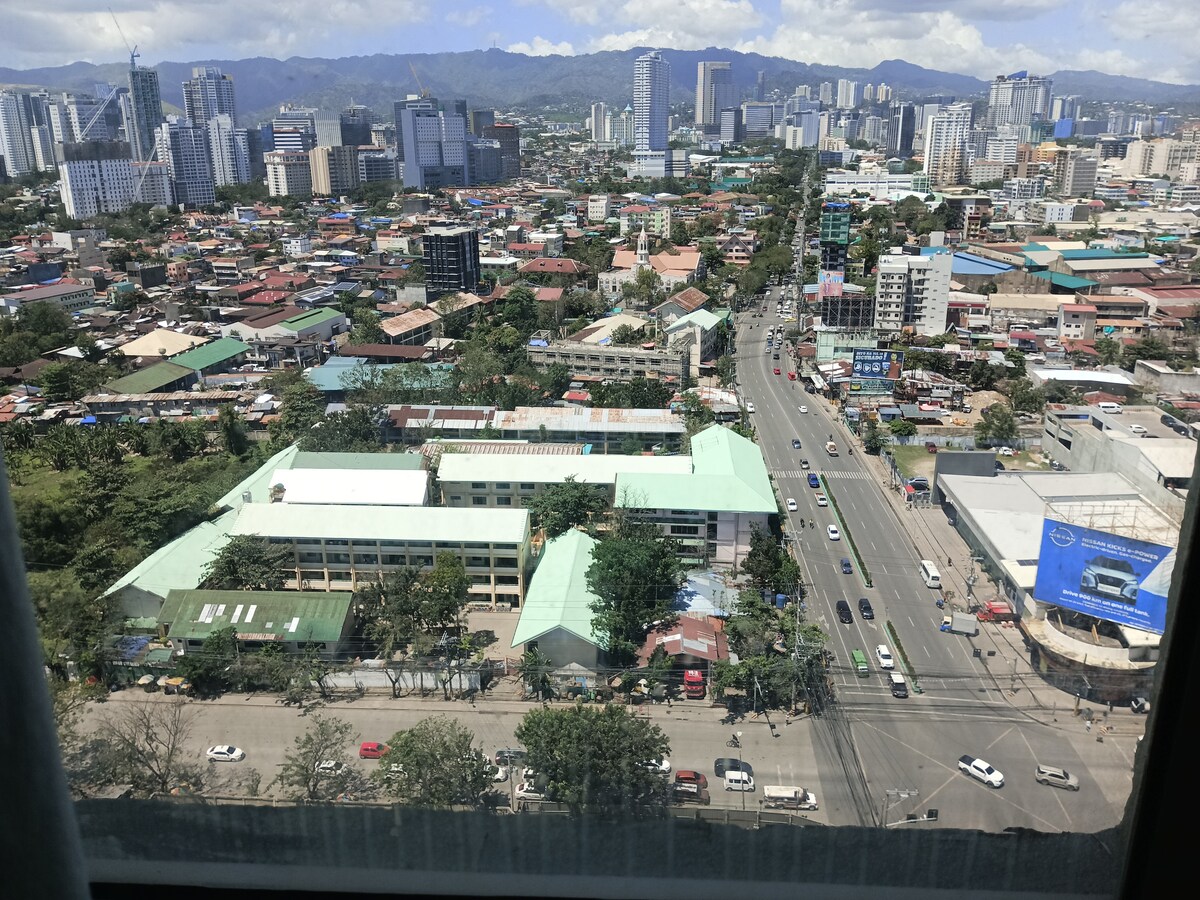 A panoramic view from the upper floor showcases a bustling cityscape, featuring a mix of high-rise buildings and low-rise structures. Green trees contrast with the urban environment, while roads are lined with vehicles. Mountains can be seen in the background beneath a partly cloudy sky.