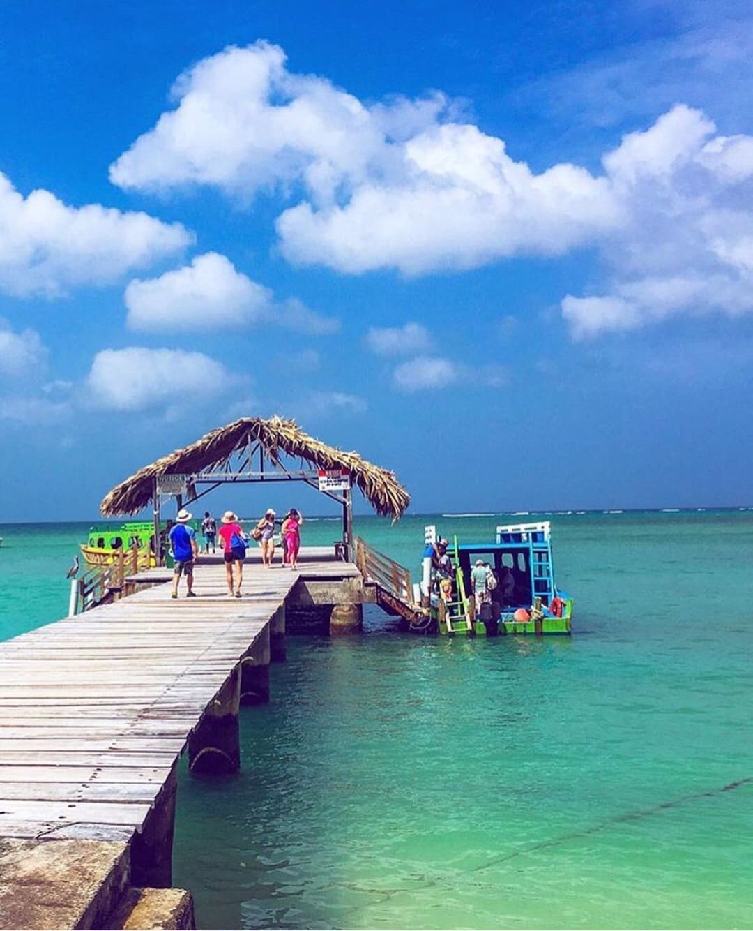 A wooden pier extends over calm turquoise waters, featuring a thatched-roof gazebo at the end. Individuals walk along the pier, while a boat is anchored nearby, creating a scene of leisure and enjoyment in a bright, sunny atmosphere.