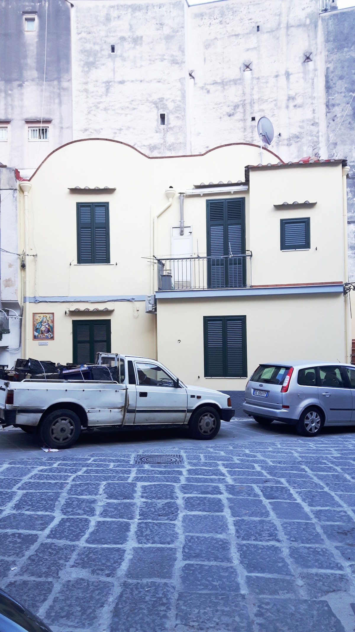 A two-story building is showcased with a distinctive curved roofline. The exterior features green shuttered windows, a balcony, and a light-colored facade. Vehicles are parked along the cobblestone street, which adds a charming atmosphere to the scene.