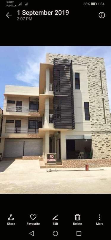 A modern three-story building features a contemporary façade with a combination of stone and smooth surfaces. Large glass windows are visible, providing ample natural light to the interior. A sign stands in the front, indicating 'No Parking,' while an open area is seen in front of the building.