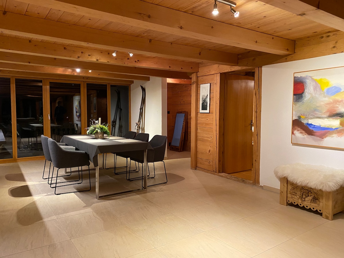 A dining area features a large rectangular table surrounded by six upholstered chairs. Natural wood beams and paneling create a warm atmosphere. A piece of abstract artwork adorns the wall beside a cozy seating area with a fur throw. Floor-to-ceiling windows allow plenty of natural light.