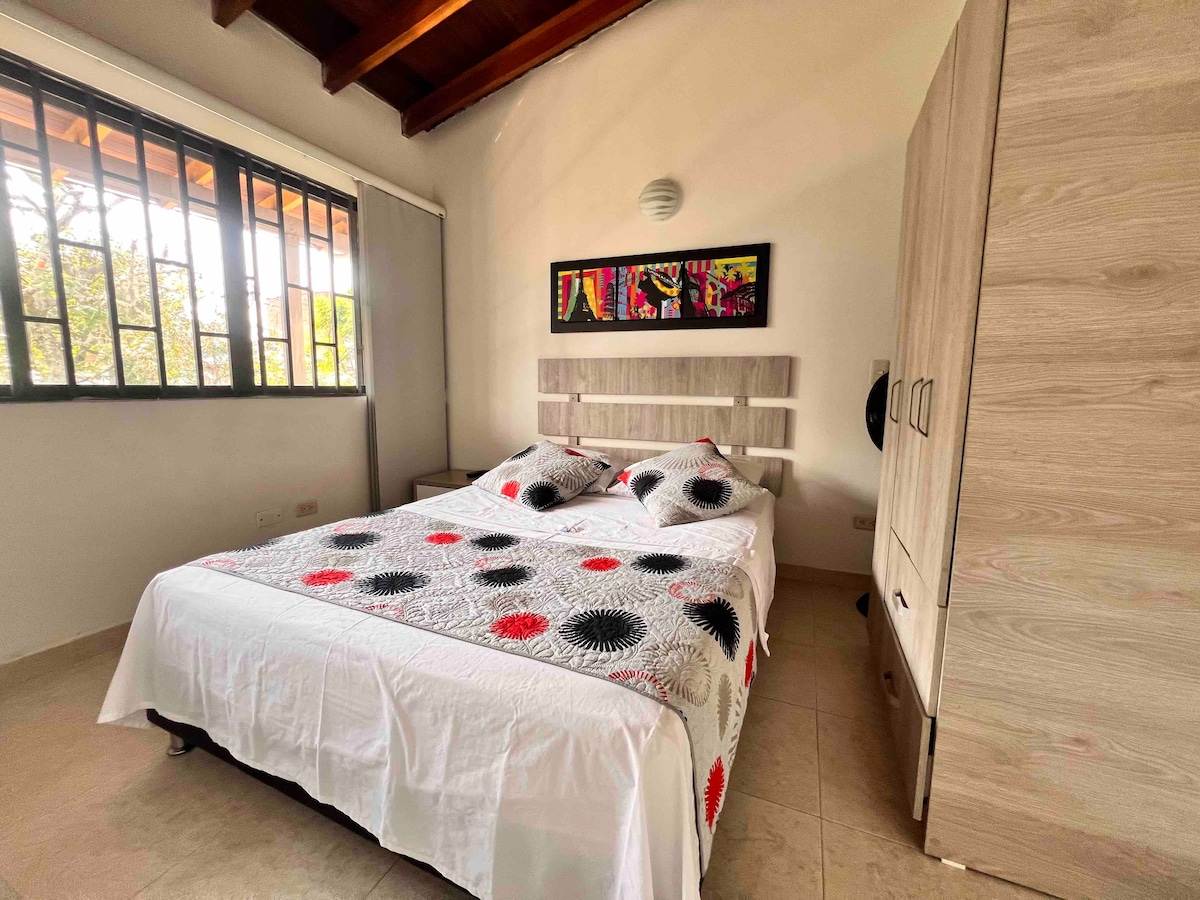 A well-lit sleeping area features a spacious bed adorned with a white and colorful patterned blanket. Artwork hangs above the headboard, and large windows provide ample natural light, framed by wooden beams and a light-colored wall cabinet.
