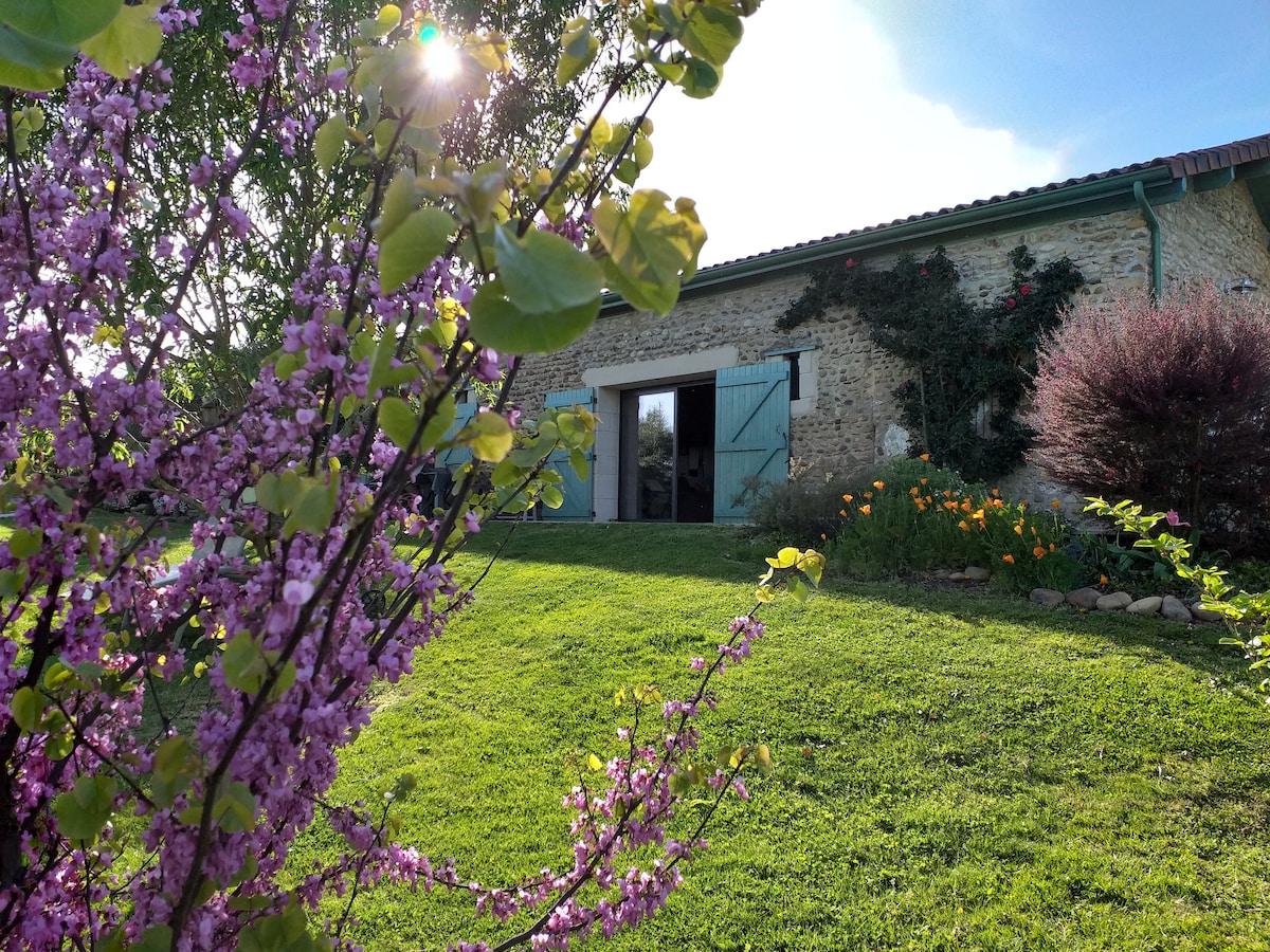 A charming stone building is set against a backdrop of greenery, with large open doors revealing a welcoming interior. A vibrant flowering tree in the foreground adds color to the scene, while the lush lawn leads up to the structure.