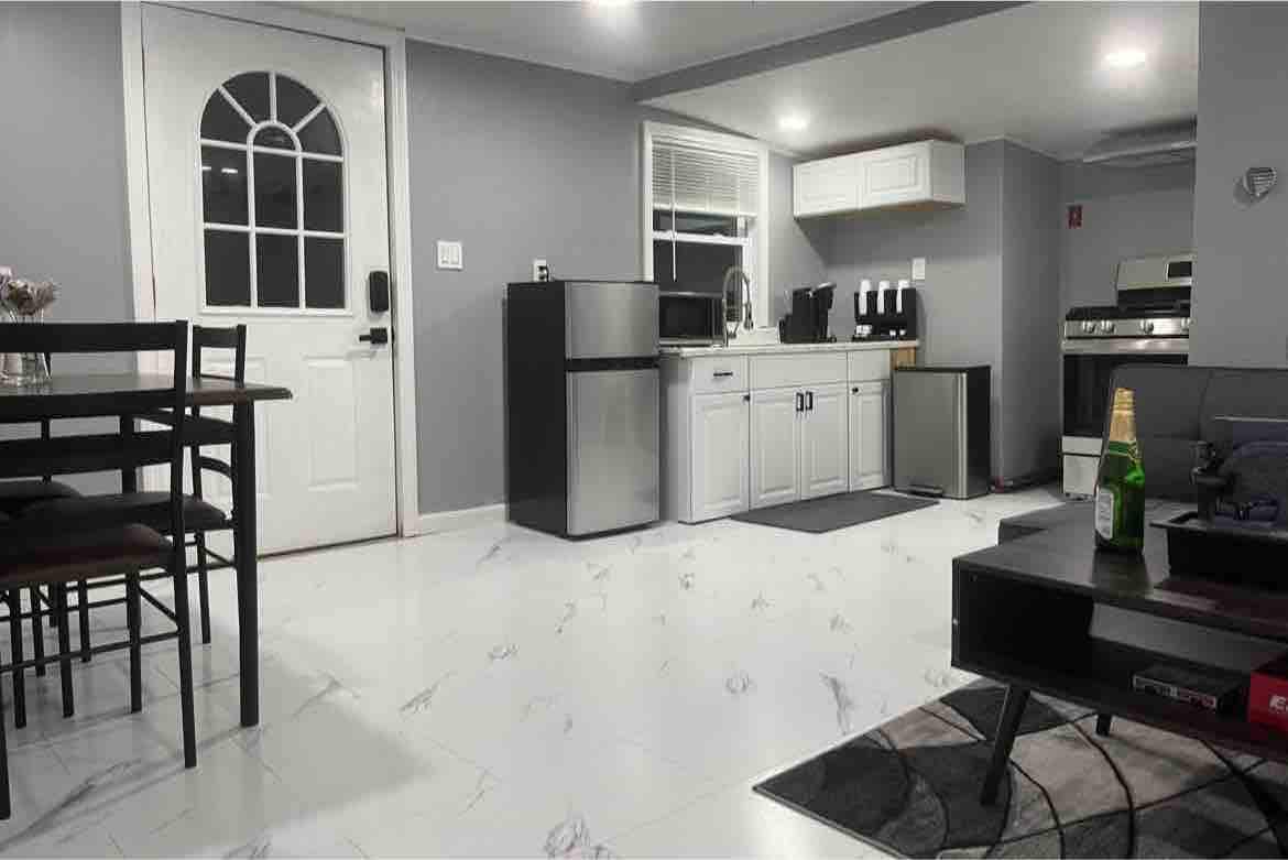 A modern kitchen area features stainless steel appliances, including a refrigerator and coffee maker. The dining table with four chairs is positioned nearby. A door with a window allows natural light to enter, highlighting the sleek marble flooring throughout the space.