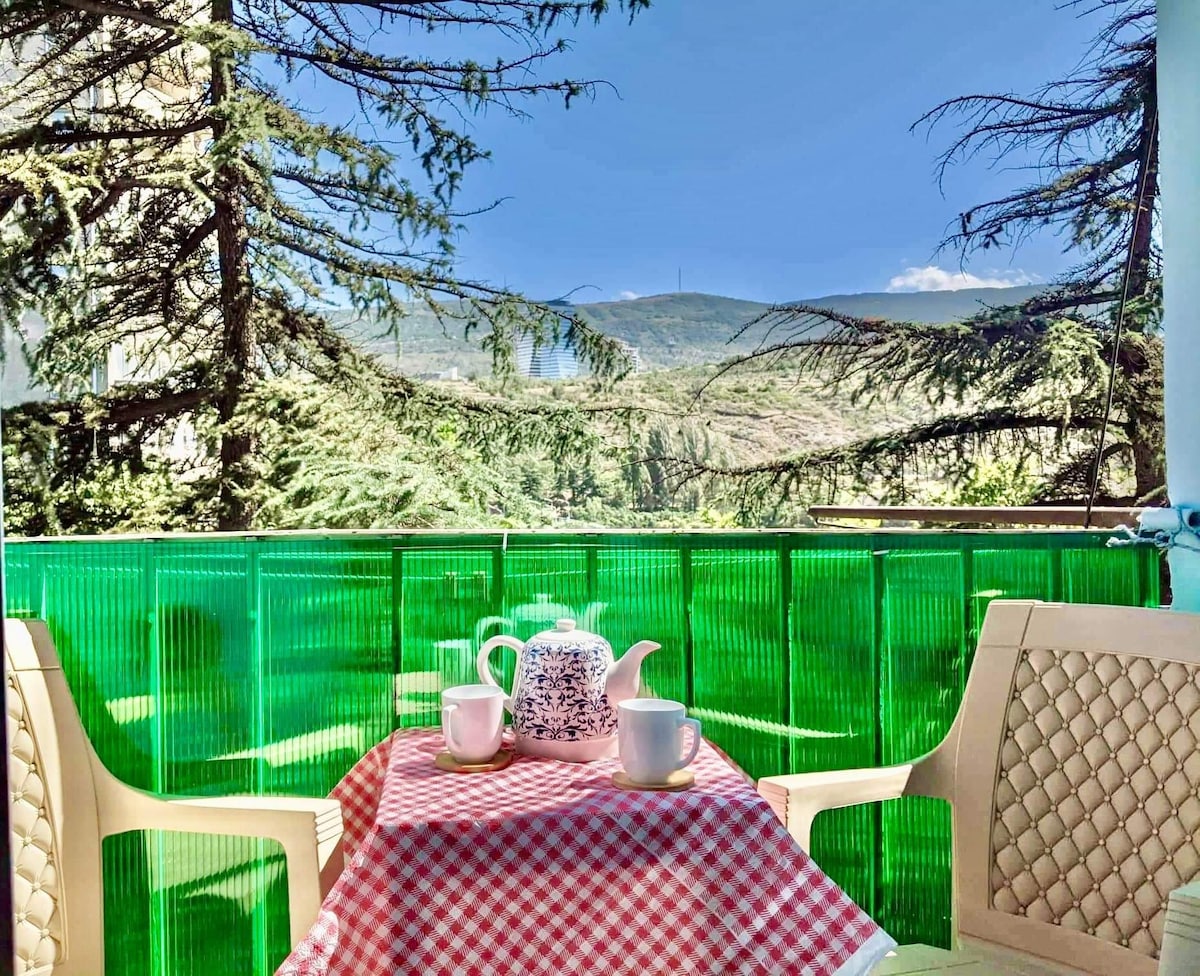 A balcony view showcases a scenic landscape, framed by green fencing and trees. A small round table is set with a checkered tablecloth, featuring a teapot and two cups. The mountains in the distance are partially visible under a clear blue sky.