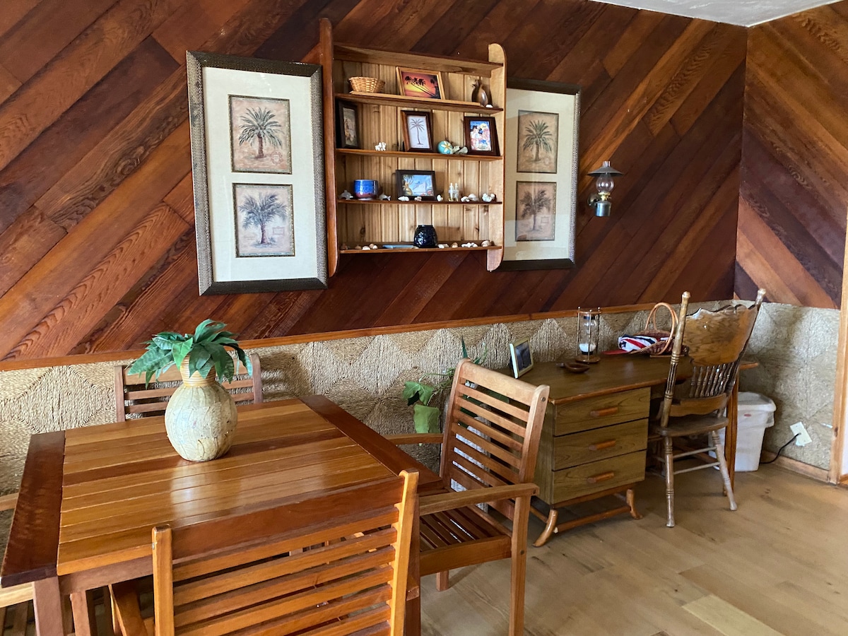 A cozy dining area features a wooden table surrounded by slatted chairs. A wooden cabinet is mounted on the wall, displaying decorative items and framed artwork. The room is enhanced by natural light filtering through nearby windows, complemented by warm wooden tones and textures.