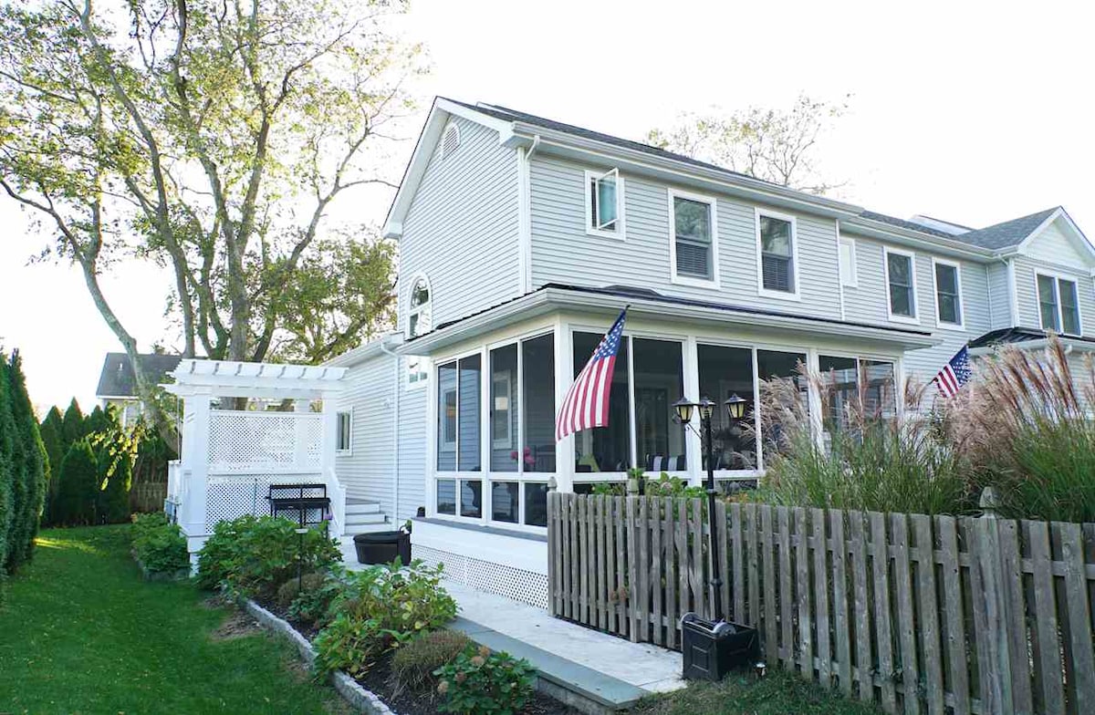 A charming townhouse exterior displays a screened porch adorned with American flags. The landscaped yard features well-maintained greenery and a rustic wooden fence. A patio area with seating is visible, inviting outdoor relaxation.