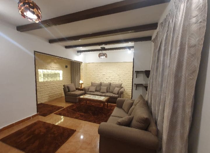A spacious living room features a comfortable arrangement of beige sofas, complemented by a wooden coffee table at the center. Soft lighting is provided by decorative pendant lights, and the textured wall adds depth to the design. Warm area rugs cover the tiled floor.