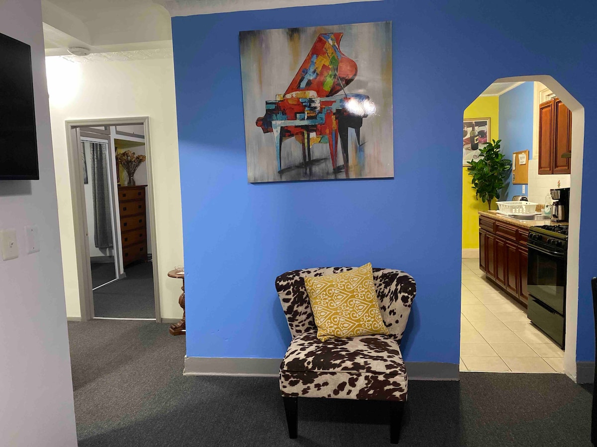 A cozy hallway showcases a brightly colored painting of a piano against a vivid blue wall. A stylish chair with a cow-print design and a golden-yellow decorative pillow is positioned nearby, inviting relaxation. The space leads toward the kitchen area, enhancing the welcoming ambiance.