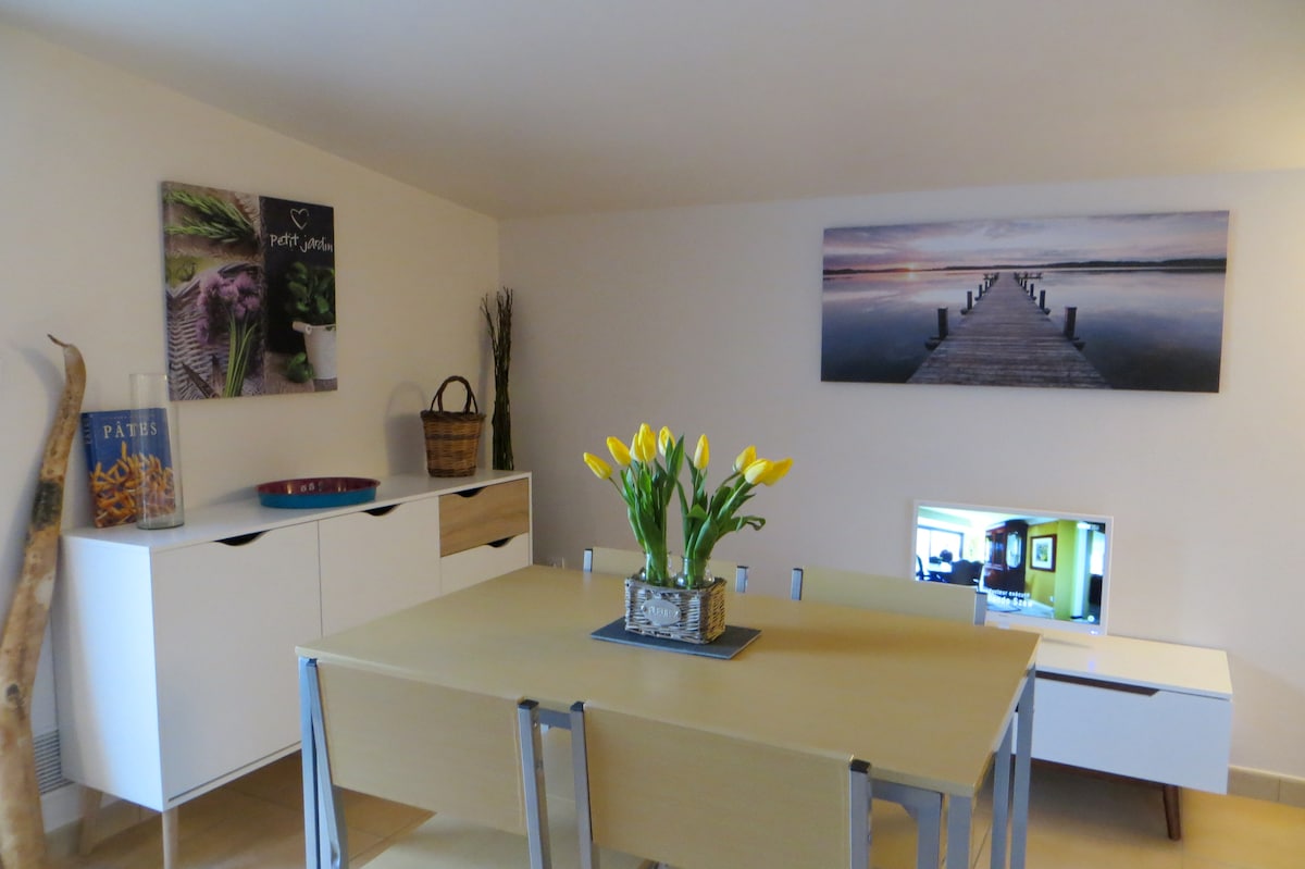 A dining area is displayed with a wooden table surrounded by metal chairs. A vase of yellow tulips serves as a centerpiece. Wall art featuring a landscape and decorative elements can be seen along with a television on a low cabinet. Natural light brightens the space.