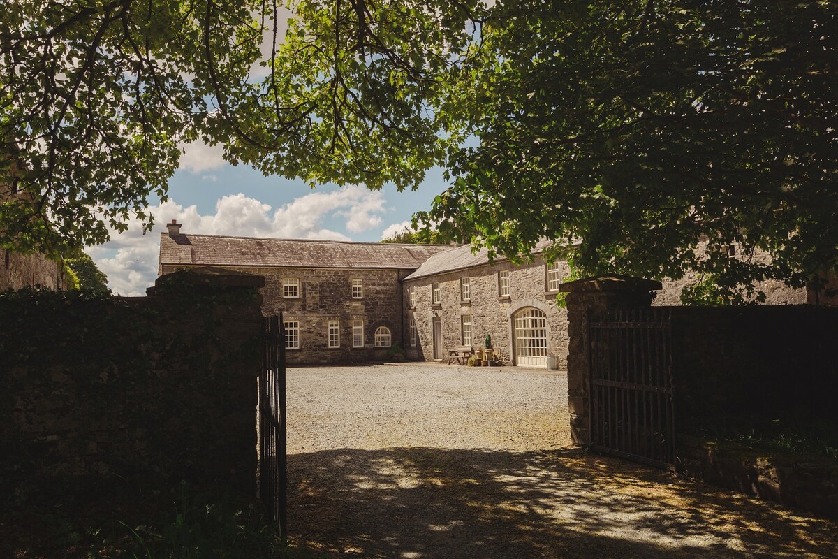 The entrance to Clonalis Estate is framed by lush green trees, revealing a gravel courtyard surrounded by elegant stone buildings. Brightly lit windows reflect the soft sunlight, while outdoor seating areas invite relaxation in the serene setting.
