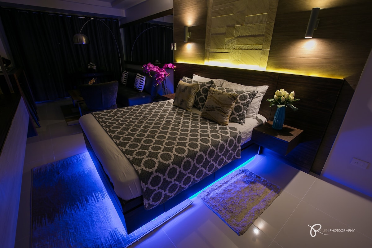 The bedroom features a spacious bed with a geometric patterned duvet and decorative pillows. Soft, ambient lighting highlights the contours of the wall behind the bed. A rug is positioned beneath the bed, complementing the overall design. Potted flowers and ambient lights add to the aesthetic appeal.