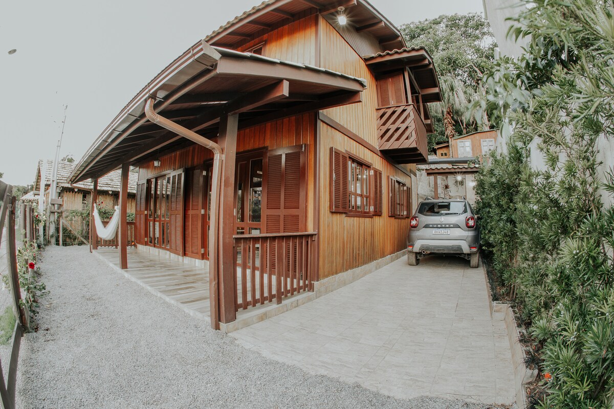 A wooden house with a charming facade is visible, featuring a covered porch that offers a relaxing space. Large windows with wooden shutters provide natural light. A small parking area next to the house is complemented by a gravel path and surrounding greenery.