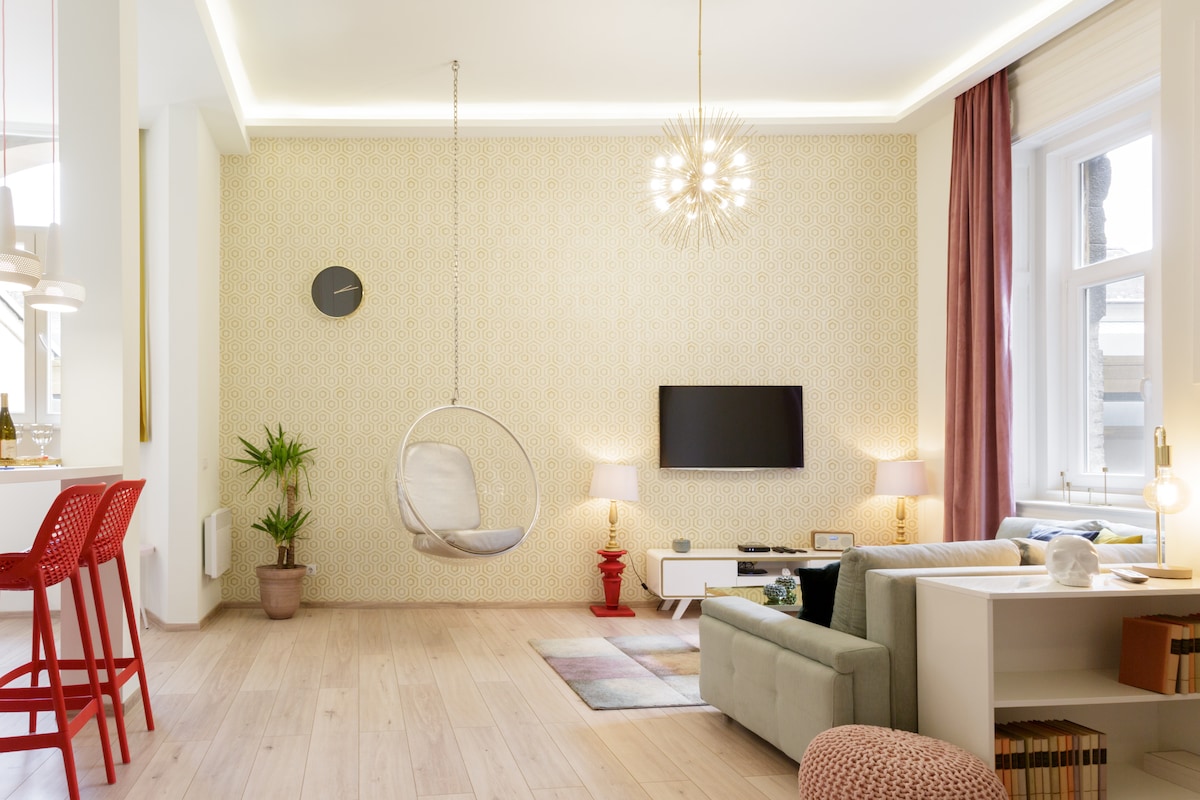 A modern living area features a hanging bubble chair suspended from the ceiling. Soft furnishings include a light green sofa and a patterned rug. Warm lighting is provided by a chandelier and table lamps, while a wall-mounted television is positioned near a decorative plant.