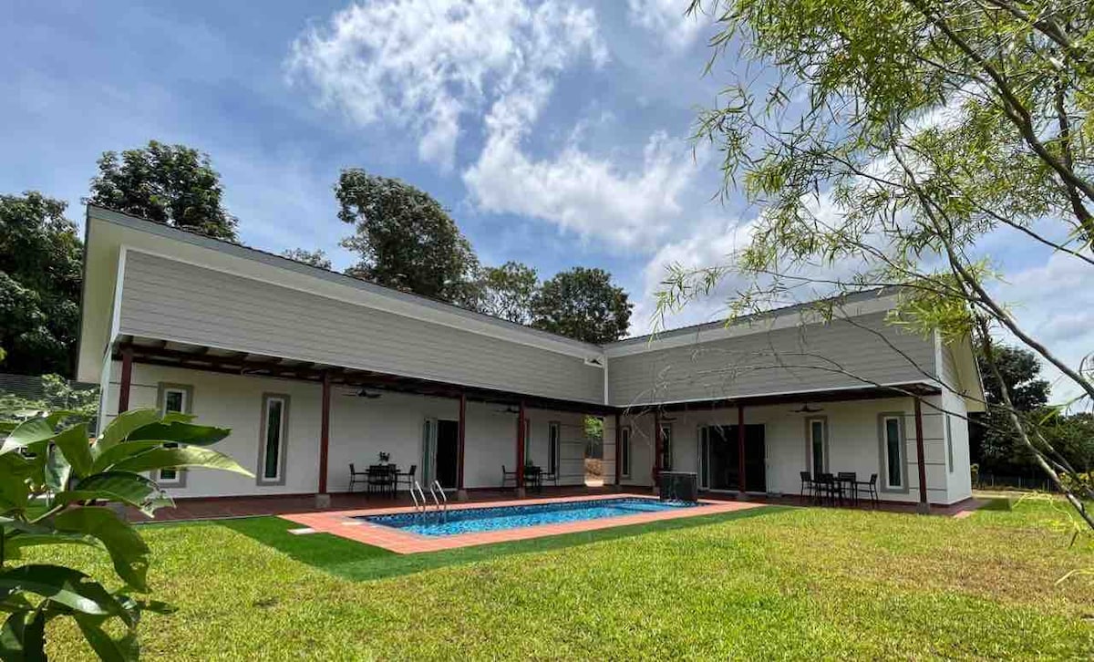 A modern villa, featuring a private pool bordered by a manicured lawn, is captured under a partly cloudy sky. Outdoor dining furniture is visible next to the pool, and large glass doors provide a connection to the indoor living space.