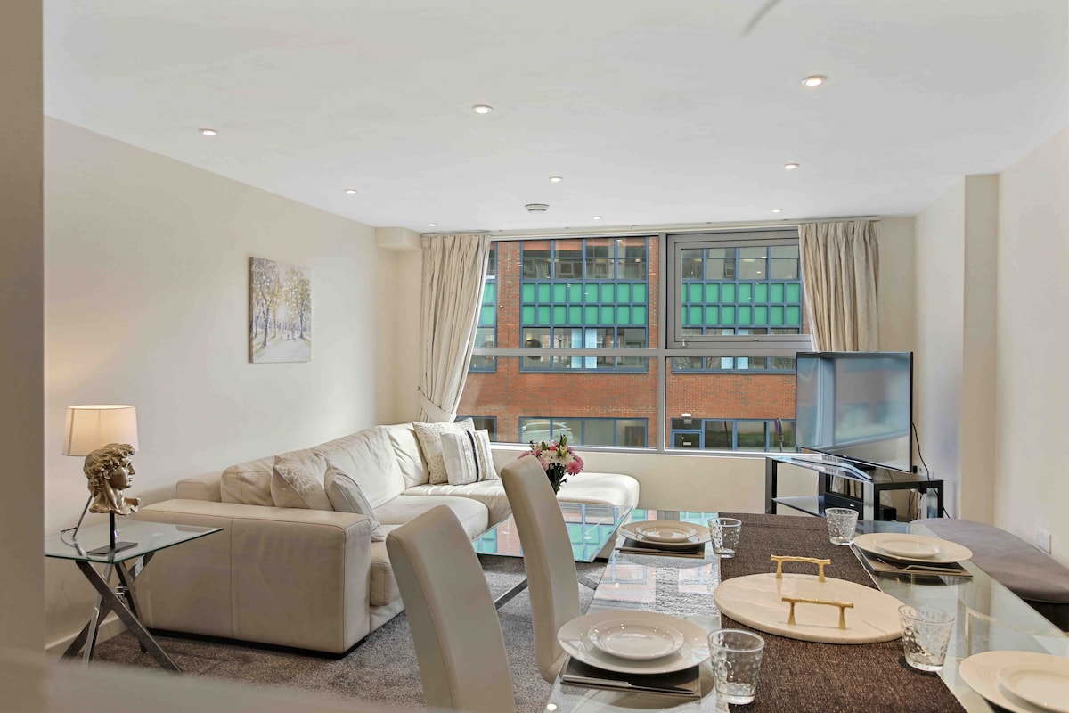 A bright living area is presented with a comfortable light-colored sofa and a glass dining table set for four. Large windows provide ample natural light, and a flat-screen TV is situated nearby, creating a welcoming environment for relaxation.