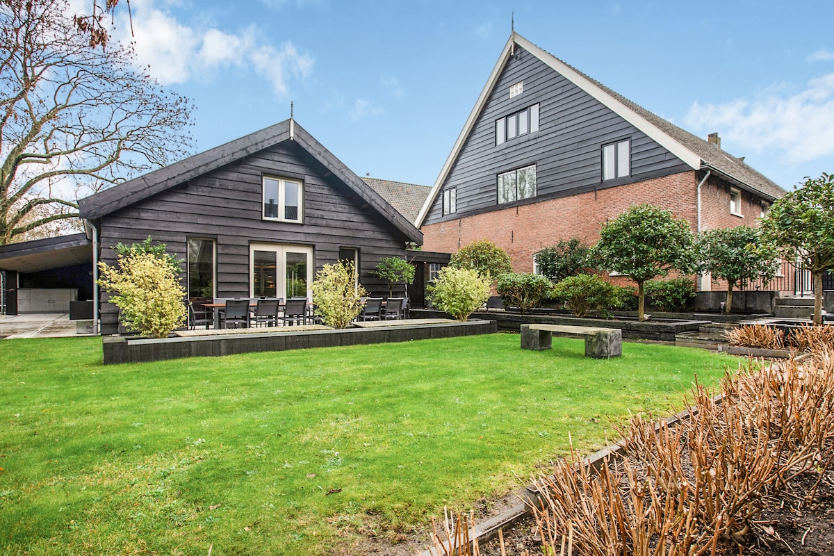 The outdoor space features a well-maintained lawn surrounded by lush greenery and shrubs. Two distinct buildings, one with a dark wood exterior and another with a brick facade, stand side by side, showcasing a blend of traditional and modern architecture.
