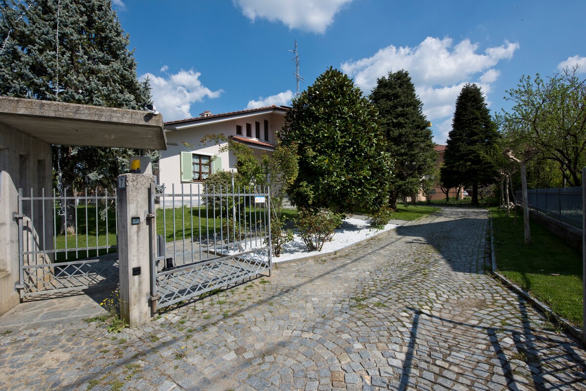 A gated entryway is visible, leading to a beautifully landscaped path lined with trees and greenery. The cobblestone driveway gently curves, offering access to the house. A well-maintained garden features shrubs and flowers, contributing to the serene outdoor environment.