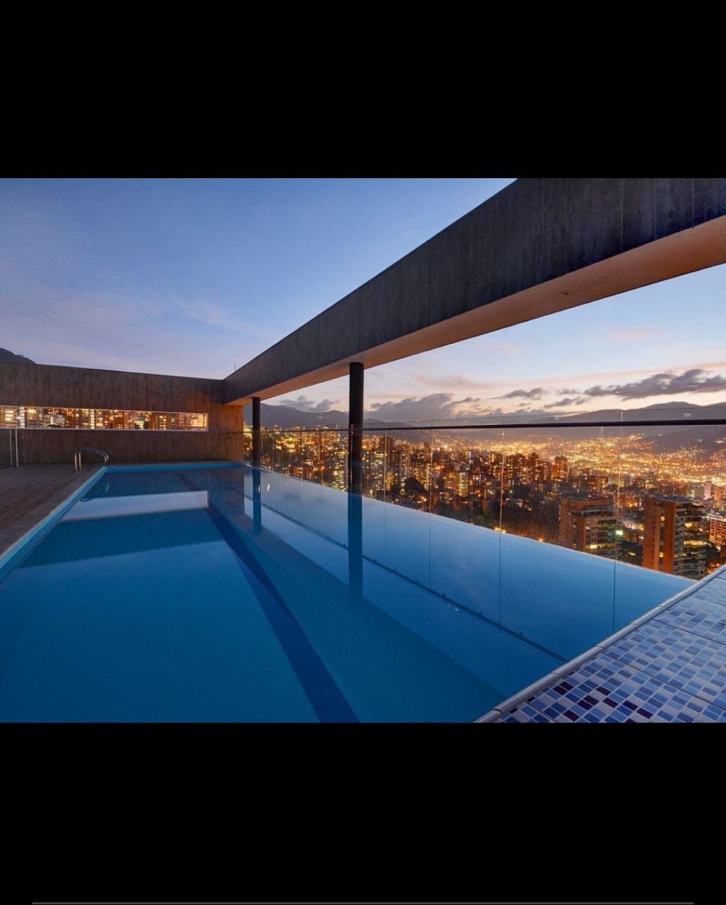 An infinity pool is showcased on a rooftop, offering stunning views of the Medellin skyline as the sun sets. Transparent glass barriers provide an unobstructed vista of the city below, creating a serene atmosphere surrounding the water.