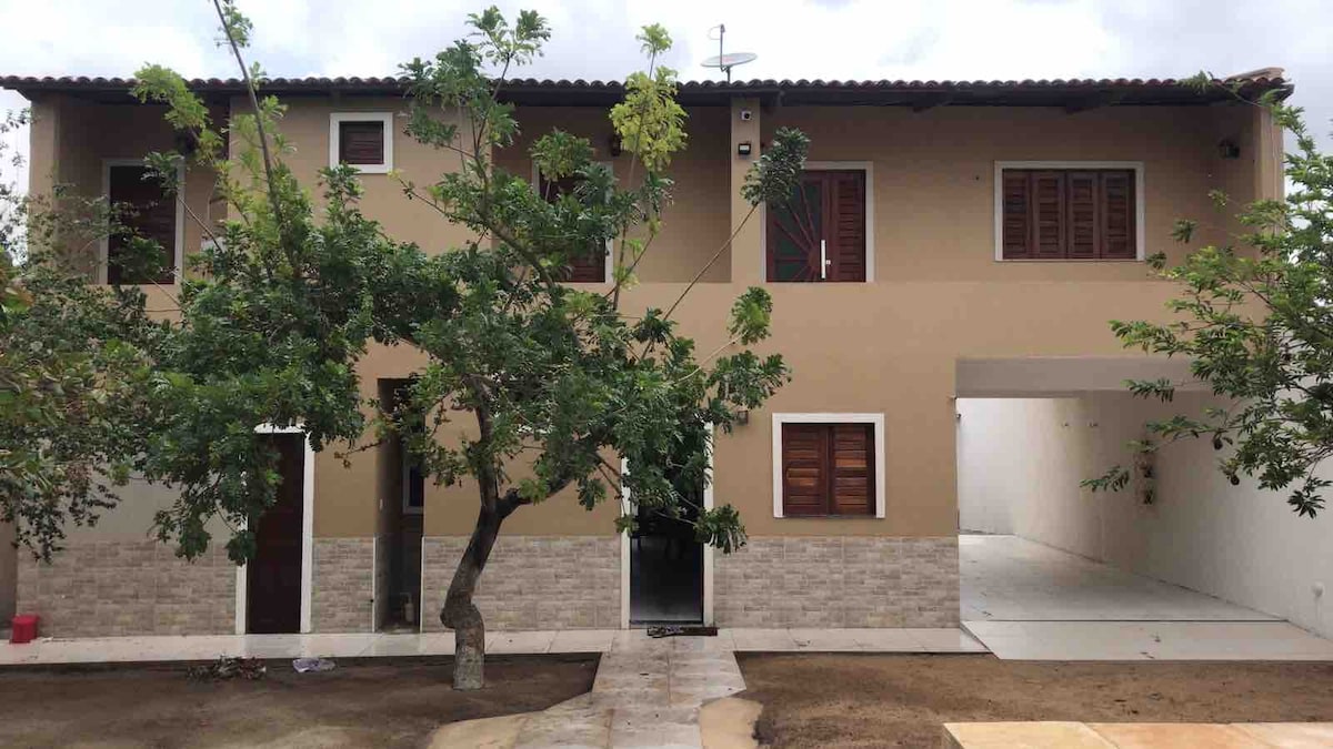 A two-story house features a combination of smooth stucco and stone facade. Windows with wooden shutters and a tree provide shade in the front yard. A paved walkway leads to the entrance, with an open space visible on the side for parking or outdoor activities.