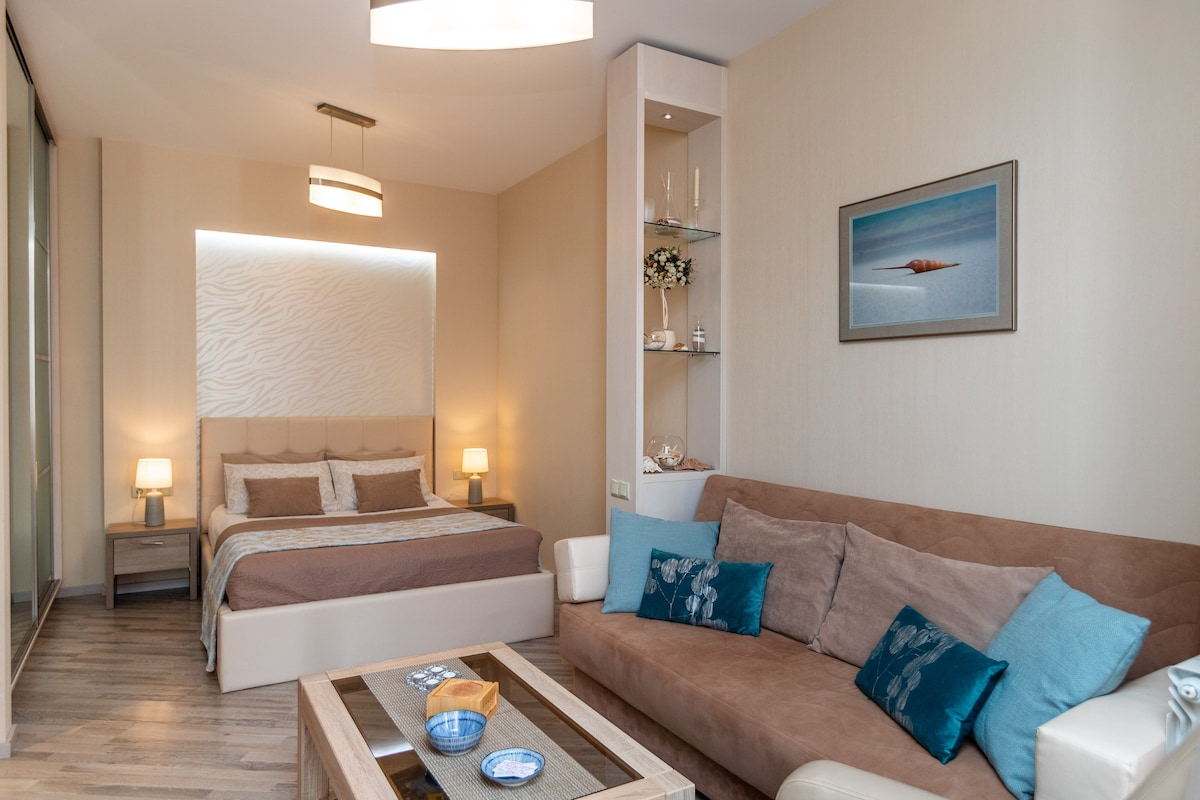 A spacious room features a comfortable bed with neutral-toned bedding. A cozy seating area includes a brown sofa adorned with blue cushions. Decorative shelves display small items, and soft light fixtures enhance the serene atmosphere.