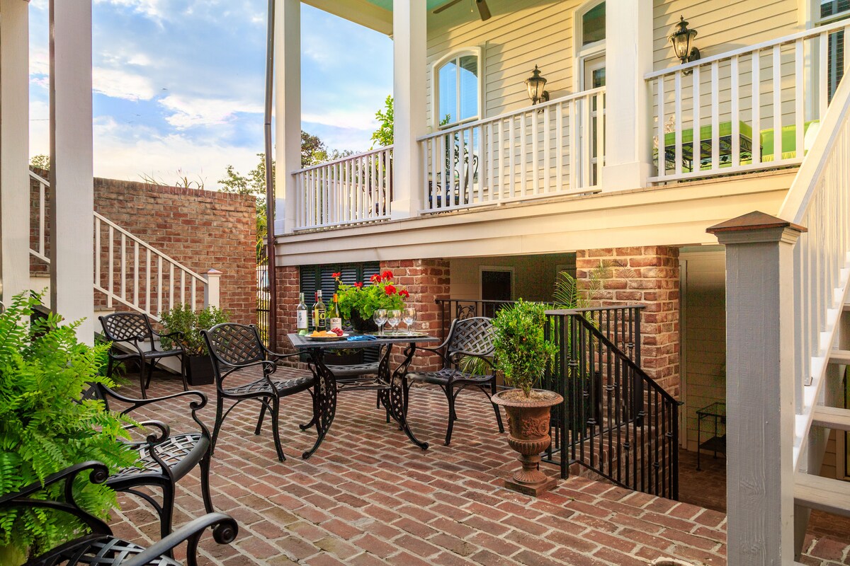 A charming outdoor courtyard is presented, featuring a brick patio surrounded by lush greenery. Several wrought iron chairs are arranged around a central table, inviting relaxation. The area's structure is accentuated by a staircase leading to a covered porch above.
