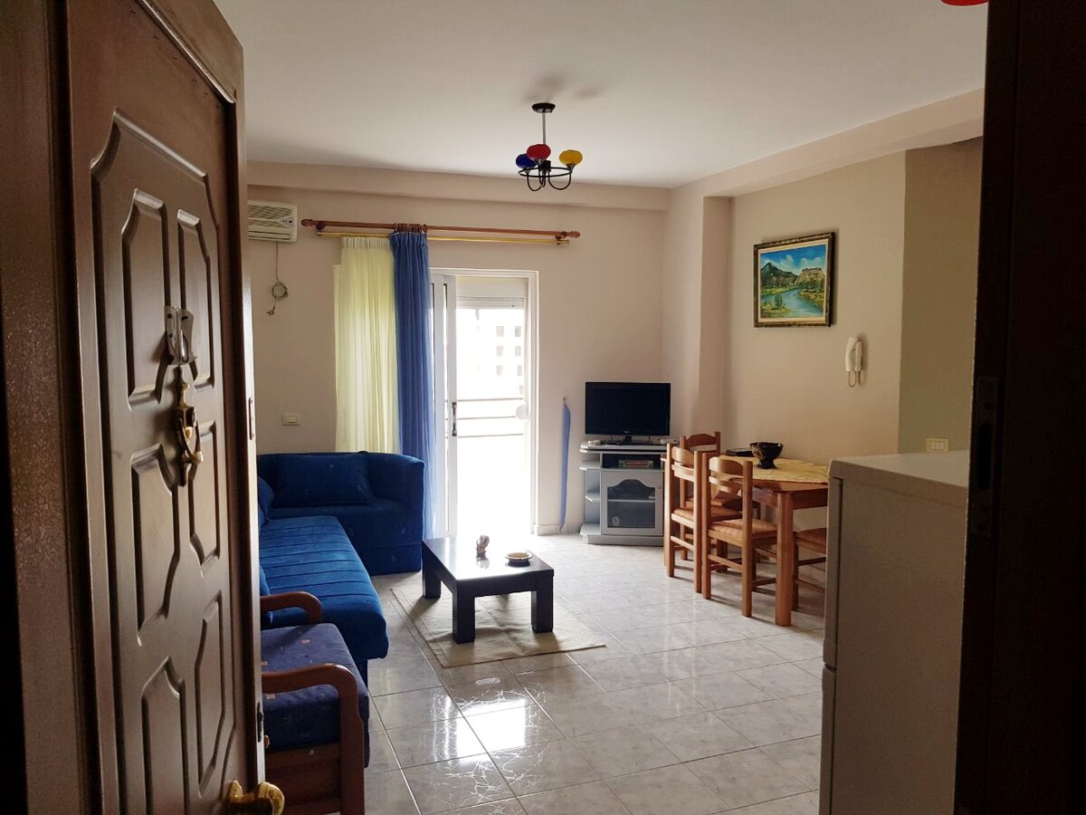 The spacious living area is characterized by a blue sofa and a coffee table, with wooden dining chairs positioned nearby. A television rests on a stand, and natural light flows through the glass door leading to the balcony. Cheerful decor is complemented by vibrant ceiling lights.