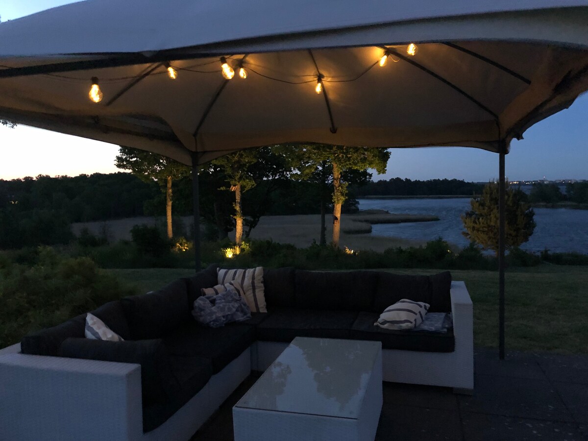 An open-air lounging area is set beneath a large shaded pavilion. A sectional sofa with cushions surrounds a glass-topped coffee table. Soft ambient lighting is provided by string lights, illuminating the space against the backdrop of a calm sea and trees visible in the distance.