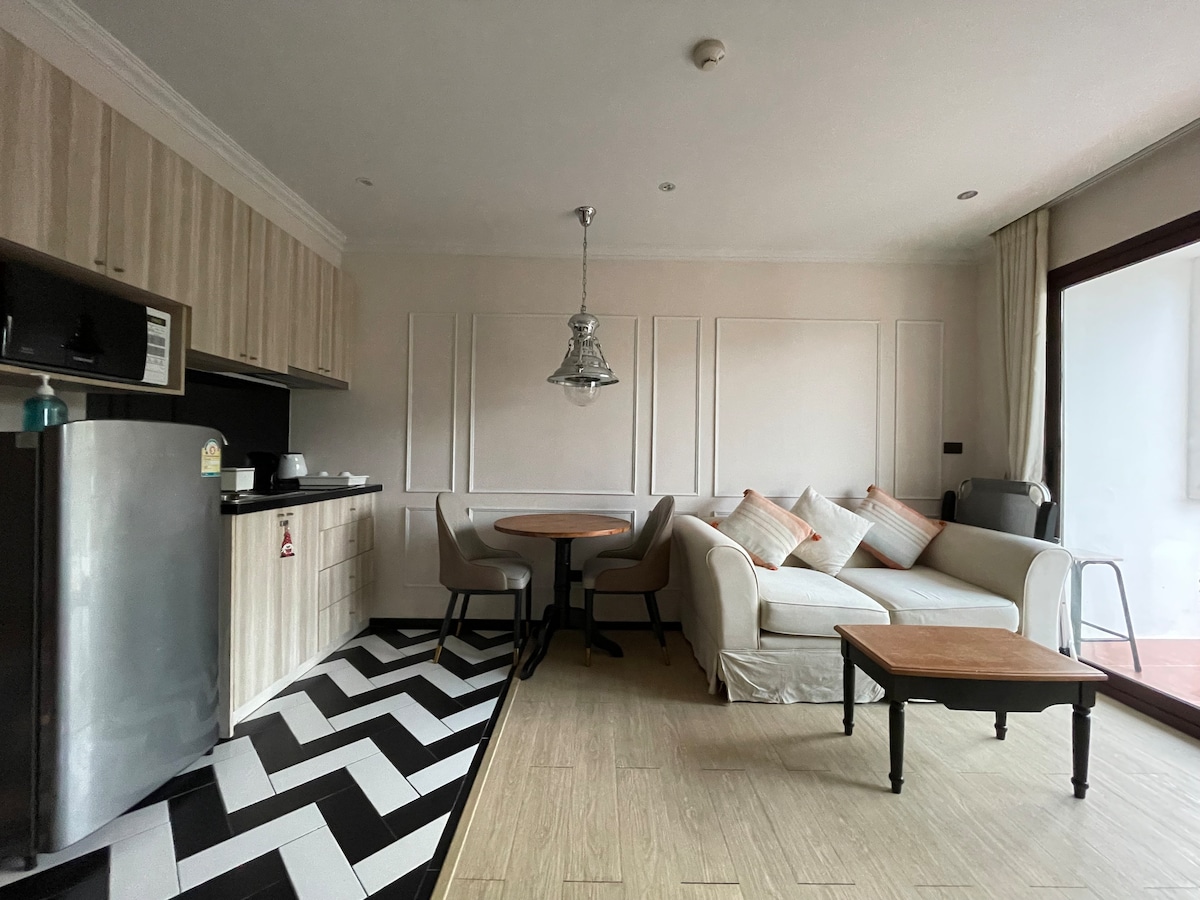 A modern living area is depicted, featuring a couch with neutral upholstery and decorative cushions. A round dining table is positioned nearby, complemented by four chairs. The kitchen area includes cabinetry and a refrigerator, while large glass doors lead to an outdoor space.
