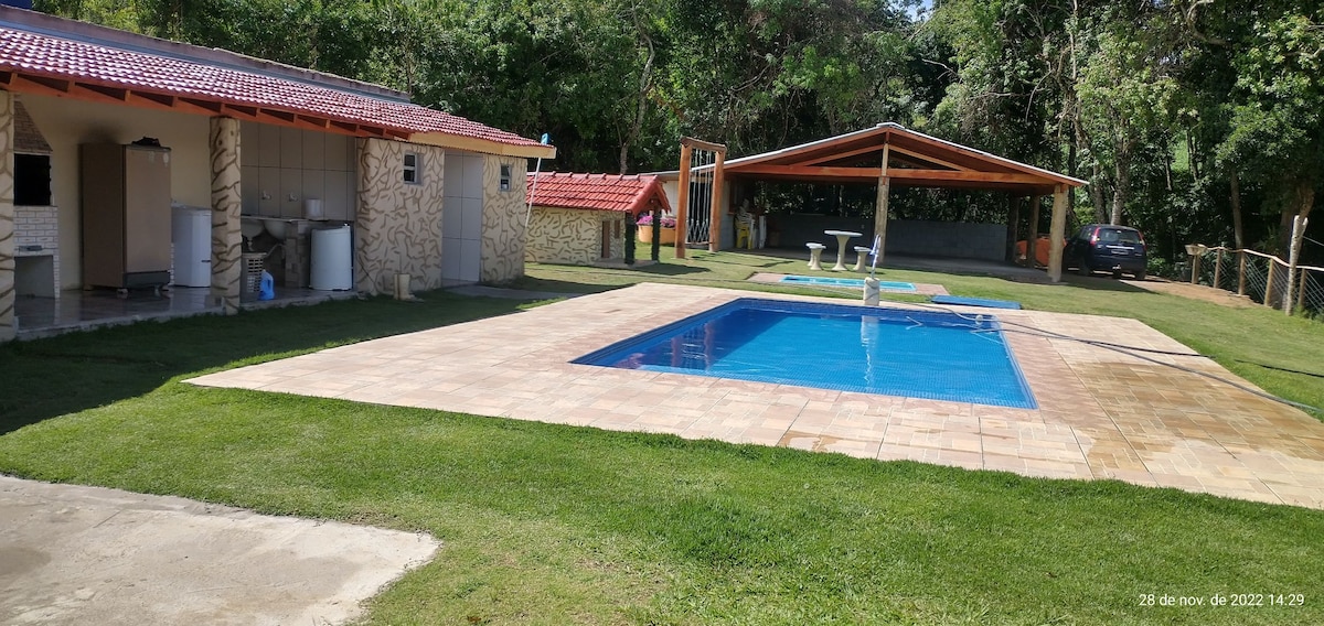 A tranquil outdoor space is displayed, featuring a well-maintained swimming pool surrounded by a paved patio. Nearby, a shaded area with picnic tables and a playground structure can be observed. Lush greenery frames the space, enhancing the serene atmosphere.