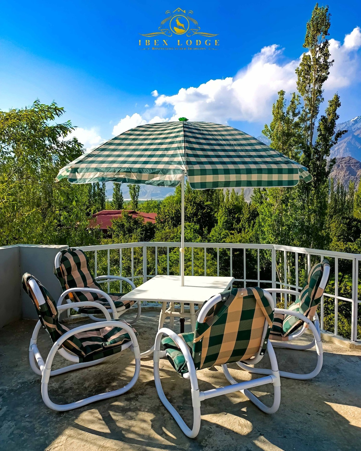 A balcony is equipped with a table surrounded by four rocking chairs. A large umbrella provides shade, and the setting features views of tall trees and distant mountains under a clear blue sky.