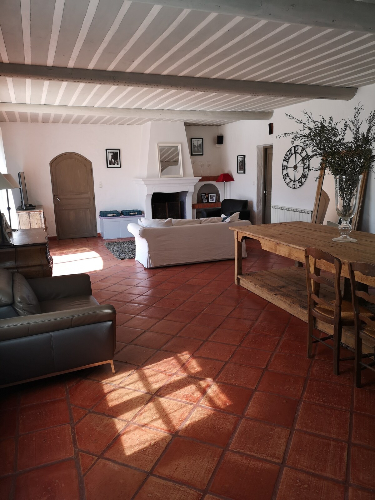 A living area features a cozy seating arrangement with a white couch and a dark leather sofa, complemented by a wooden dining table. A fireplace serves as a focal point, with decorative elements like a large clock and artwork on the walls. Natural light illuminates the space.