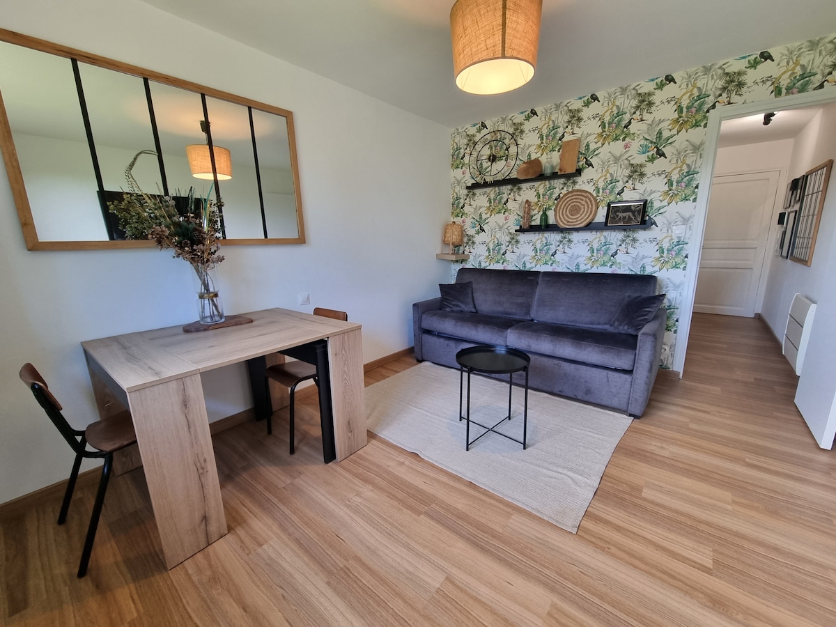 A modern living area features a gray sofa facing a small coffee table, with a wooden dining table and two chairs to the side. A botanical-themed accent wall adds visual interest, complemented by a large mirror and decorative pieces on the shelves.