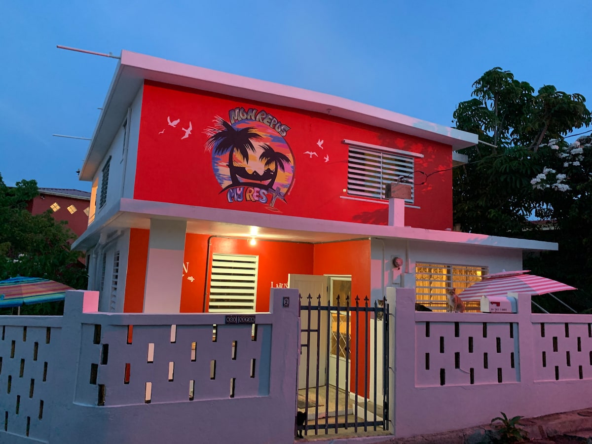 The exterior of the studio is highlighted by its vibrant red walls adorned with tropical motifs. A gated entrance leads to the inviting front area, while colorful umbrellas provide shade. Soft evening light casts a warm glow, enhancing the welcoming atmosphere of the property.