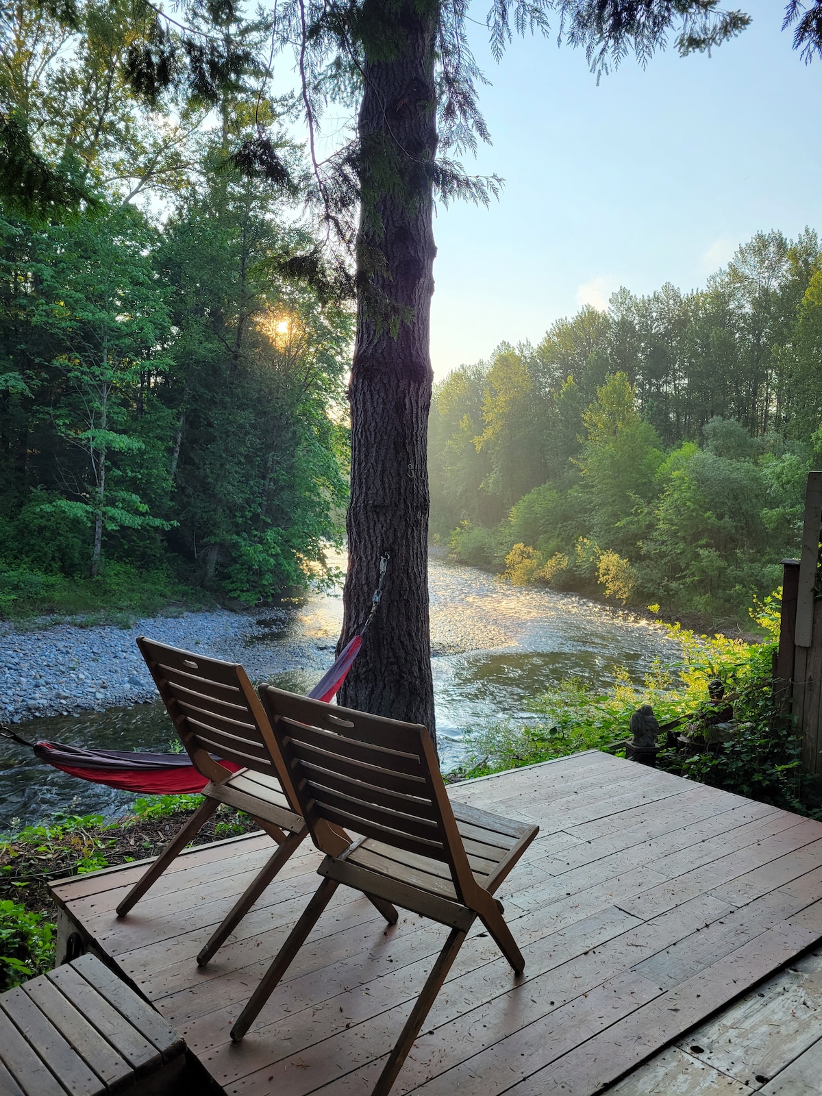 A wooden deck features two slatted chairs, positioned to face the tranquil South Fork of the Snoqualmie River. Lush greenery surrounds the area, and sunlight filters through the trees, creating a serene atmosphere by the water.