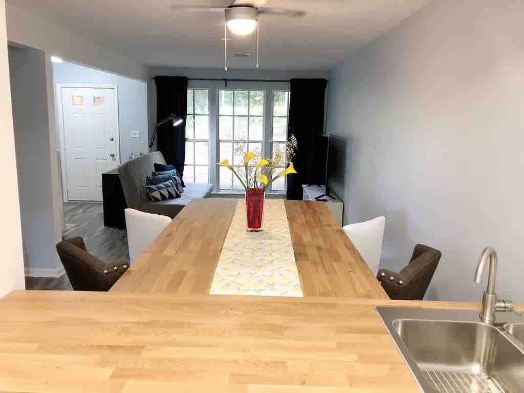 A spacious dining area features a wooden table set with a vase of flowers at its center. Chairs are positioned around the table, and a view into the living room is visible in the background, showcasing large glass doors leading to an outdoor space.