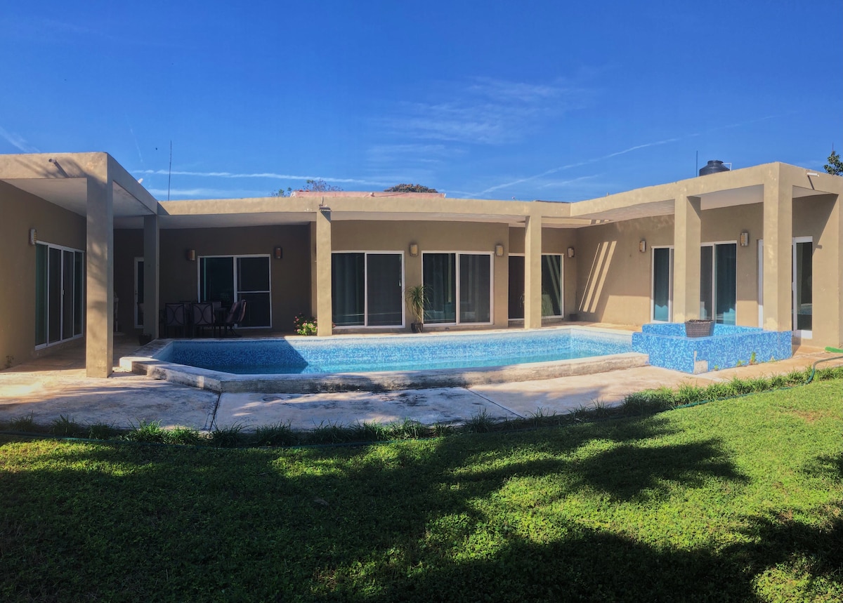 The exterior of a modern villa is depicted, featuring a large swimming pool surrounded by a spacious patio. Floor-to-ceiling windows enhance the natural light, and lush green grass adds a serene touch to the outdoor area.