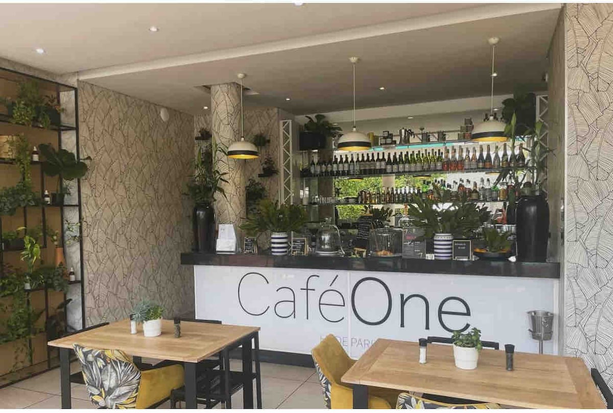 A well-lit café area features a sleek counter adorned with a variety of plants. Shelves behind the counter display an organized selection of beverages. Small tables with stylish chairs are arranged for seating, creating a relaxed environment. Natural light brightens the space.