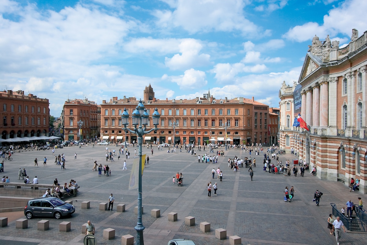 A spacious public square is seen, featuring a mix of people walking and sitting on benches. Historic buildings with red brick facades surround the square, complemented by a blue sky with scattered clouds. Streetlamps and benches add to the open ambiance.