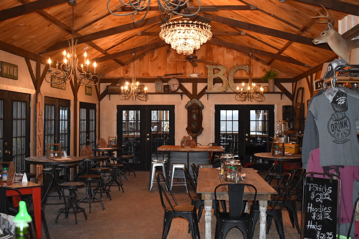 An inviting communal space is presented with a wooden ceiling and rustic decor. Multiple wooden tables and metal chairs are arranged throughout. A large chandelier hangs above, complemented by wall-mounted lighting. Two double doors offer access to the outside, and a bar area is visible in the background.