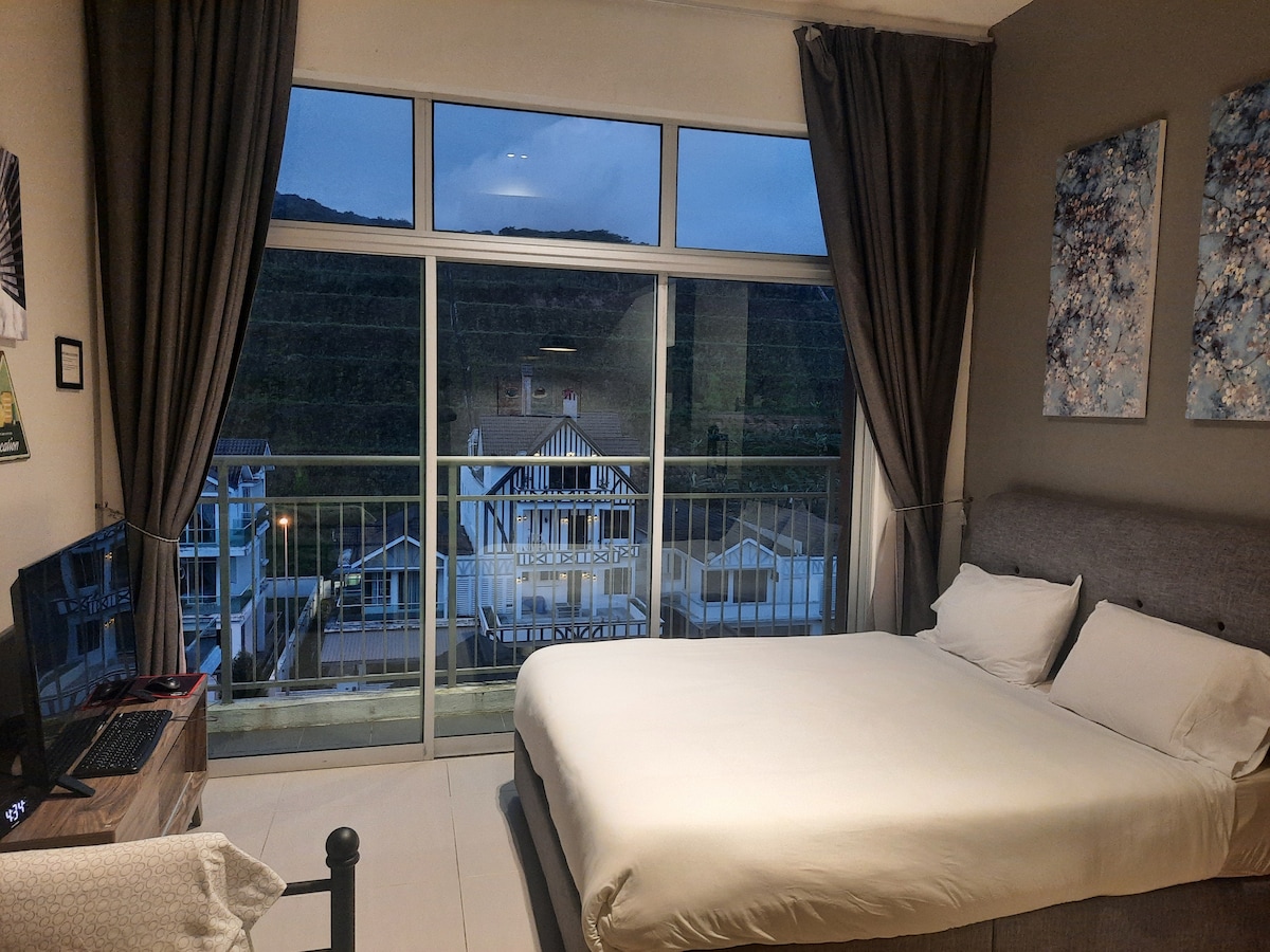 A comfortable sleeping area is featured with a spacious bed positioned near large glass doors. The view outside includes distant hills and nearby buildings, with soft drapes framing the window. A television is mounted on a cabinet beside the bed.