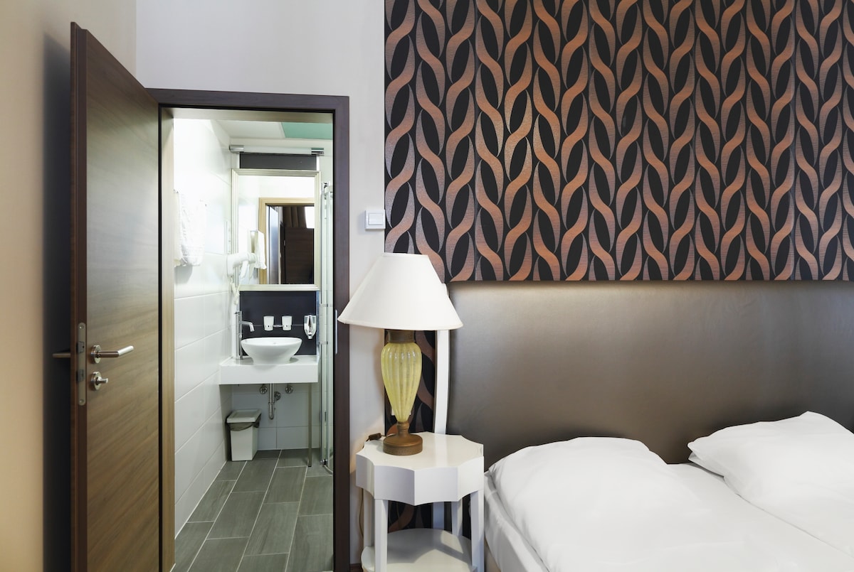 A stylish studio is displayed with a patterned wall featuring waves in shades of brown and cream. A spacious bed is positioned in the foreground, while a bathroom is accessible through an open door, showcasing modern fixtures and bright lighting.