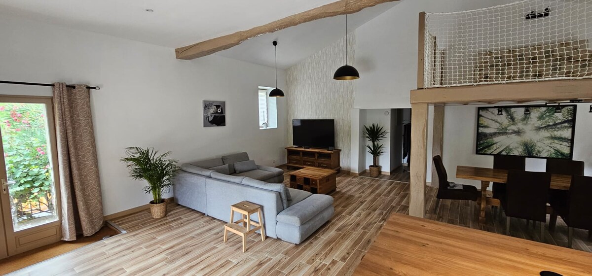 An open and airy living space features a light grey sectional sofa arranged around a wooden coffee table. A large television is positioned on a wooden console. Natural light enters through windows, enhancing the neutral tones of the decor and the warm wood flooring.